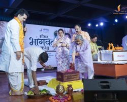 Group of people on a stage performing a Hindu ritual with fruits, flowers, and brass lamps; a woman in lavender sari assists as others look on, backdrop shows a banner and decorations.