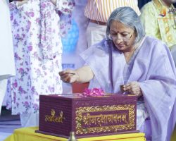 Elderly woman in a lavender sari performs a ritual, cutting with a small knife beside an ornate box. Behind her, people in colorful clothing watch.