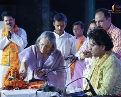 Senior woman in a lavender sari lights or arranges ritual items at a ceremony while others in traditional attire observe on a stage with marigold garlands.