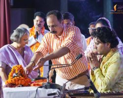 Group of people in traditional attire performing a ritual on a stage, with marigold garlands and microphones nearby.