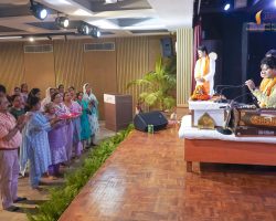 Audience in traditional Indian attire standing and clapping for a performer on stage with garlands and a harmonium nearby.