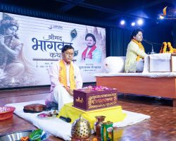 A man in traditional white attire sits cross‑legged on a stage, wearing an orange scarf, surrounded by ceremonial items and flower garlands, while another man speaks at a microphone on the right, with a large printed backdrop behind them.