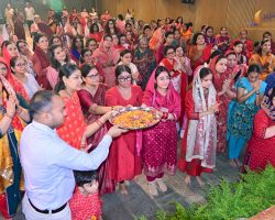 Large group of Indian women in bright sarees and salwar kameez taking part in a flower-petal ceremony in a hall. A man in a light shirt holds a tray of petals in the foreground.