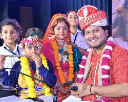 Group of children and a man in festive Indian wedding outfits on stage, with the bride at center wearing red and orange garlands.