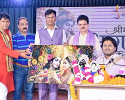 Group of men on a stage presenting a colorful painting of Hindu deities, with marigold garlands in the foreground.