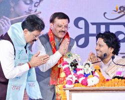 Three men in traditional Indian attire bow with folded hands on a stage during a ceremony, surrounded by flower garlands and a decorated idol centerpiece.