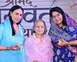 Three smiling Indian women in colorful outfits pose together, each holding a sweet treat, at an event backdrop.