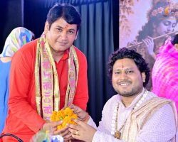 Two men in traditional Indian attire exchange a marigold bouquet during a ceremony; one in a red kurta with a decorative scarf, the other in lavender attire with beads on a backdrop stage.